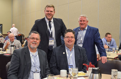 Photo of 4 WHLA Board & Committee members at our annual conference & trade show. Four men in business professional suites, two sitting at a round table, and two standing behind them.