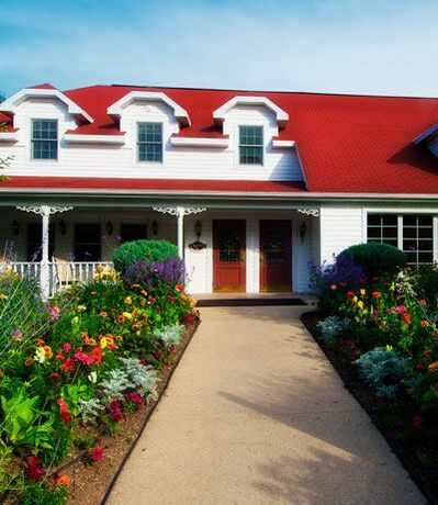 Exterior photo of High Point Inn in the springtime. Sidewalk leading up to the front red doors. Path lined with blossoming flowers. Bright red roof on a very blue sky day.