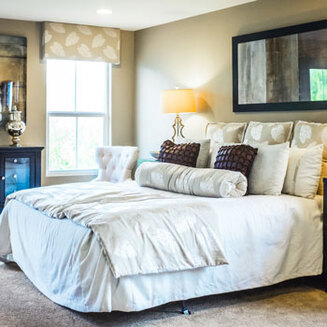 Side profile of a bed with a white comforter, tan blankets, lots of square throw pillows that are tan and deep purple, and one tubular pillow in the front.