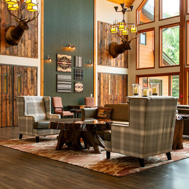 Photo of a rustic interior of a hotel lobby area. Lots of wood, large windows, green plaid chairs, green walls, eclectic wooden tables, and two deer heads mounted on the back walls.