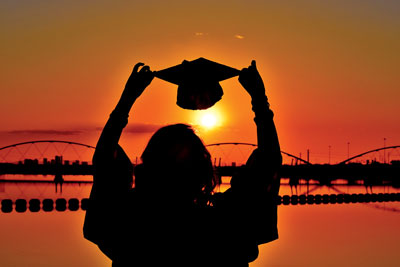 Silhouette of a student holding a graduation cap in the air during sunset over the water, with a bridge in the background