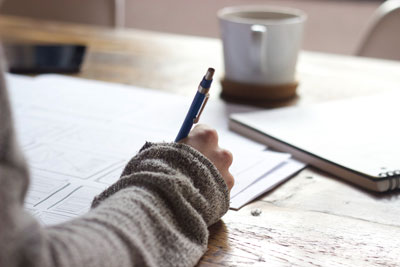 Picture of someone with a sweater, writing on a piece of paper, with a coffee cup blurred in the background.